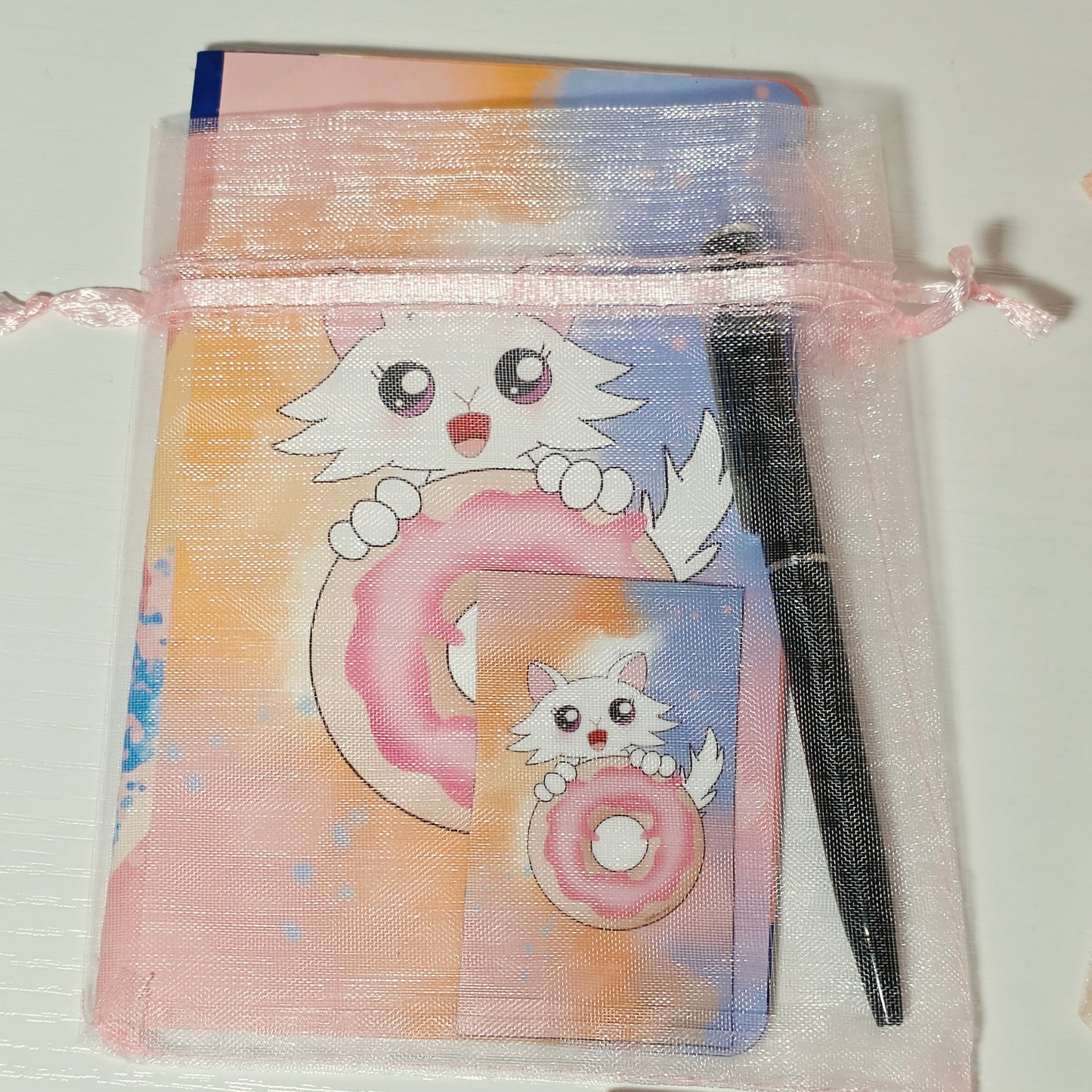 Small notepad with Daisy the Cat holding a donut on the cover. Mini pen and magnet included inside a pink mesh bag with pink ribbon